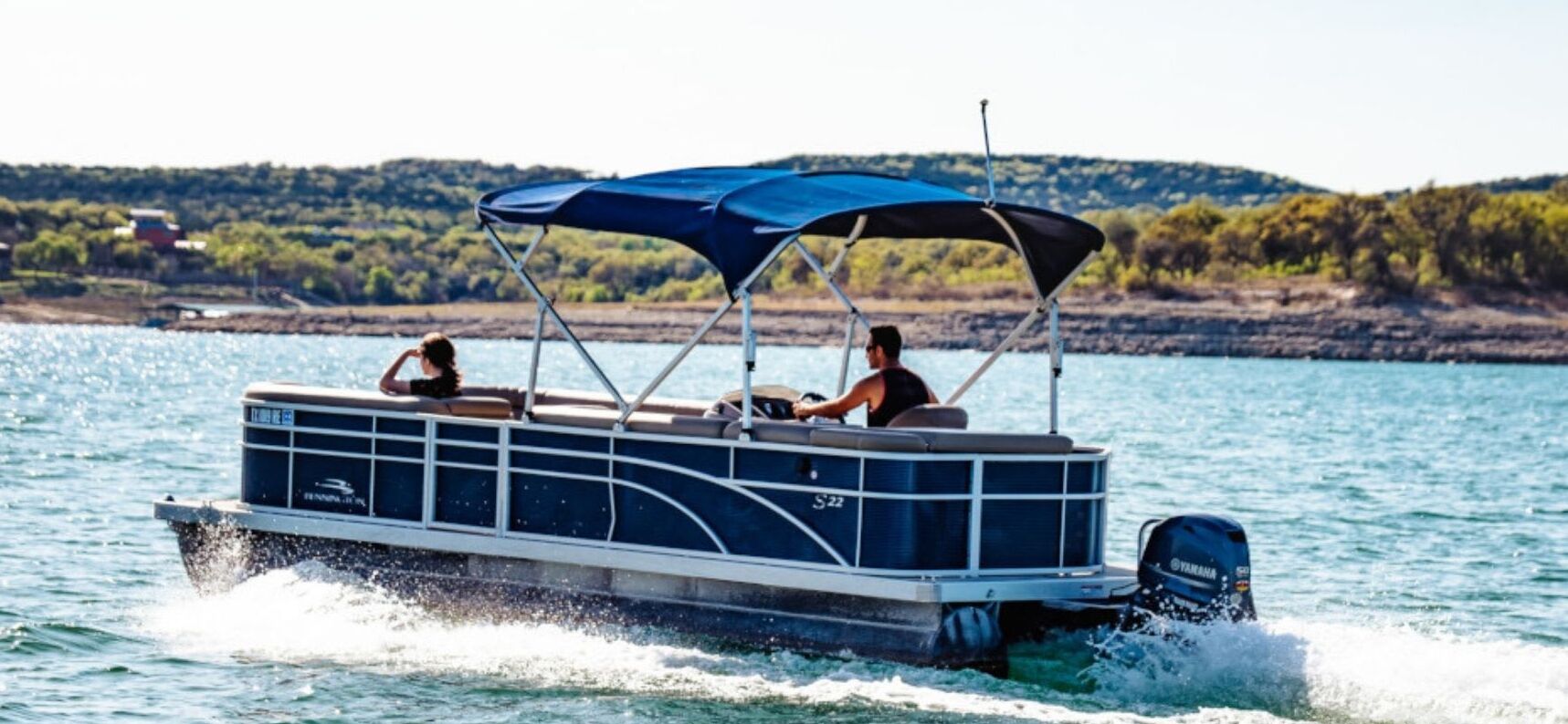 A pontoon boat with a canopy, two people, and a shoreline in the background.