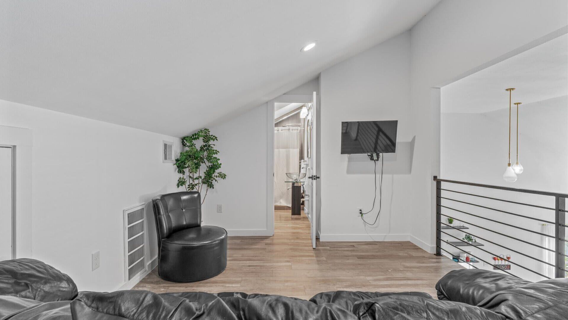 Un salón moderno con un sofá y una butaca de cuero negro, una planta en maceta, un televisor montado en la pared y una escalera con barandilla.