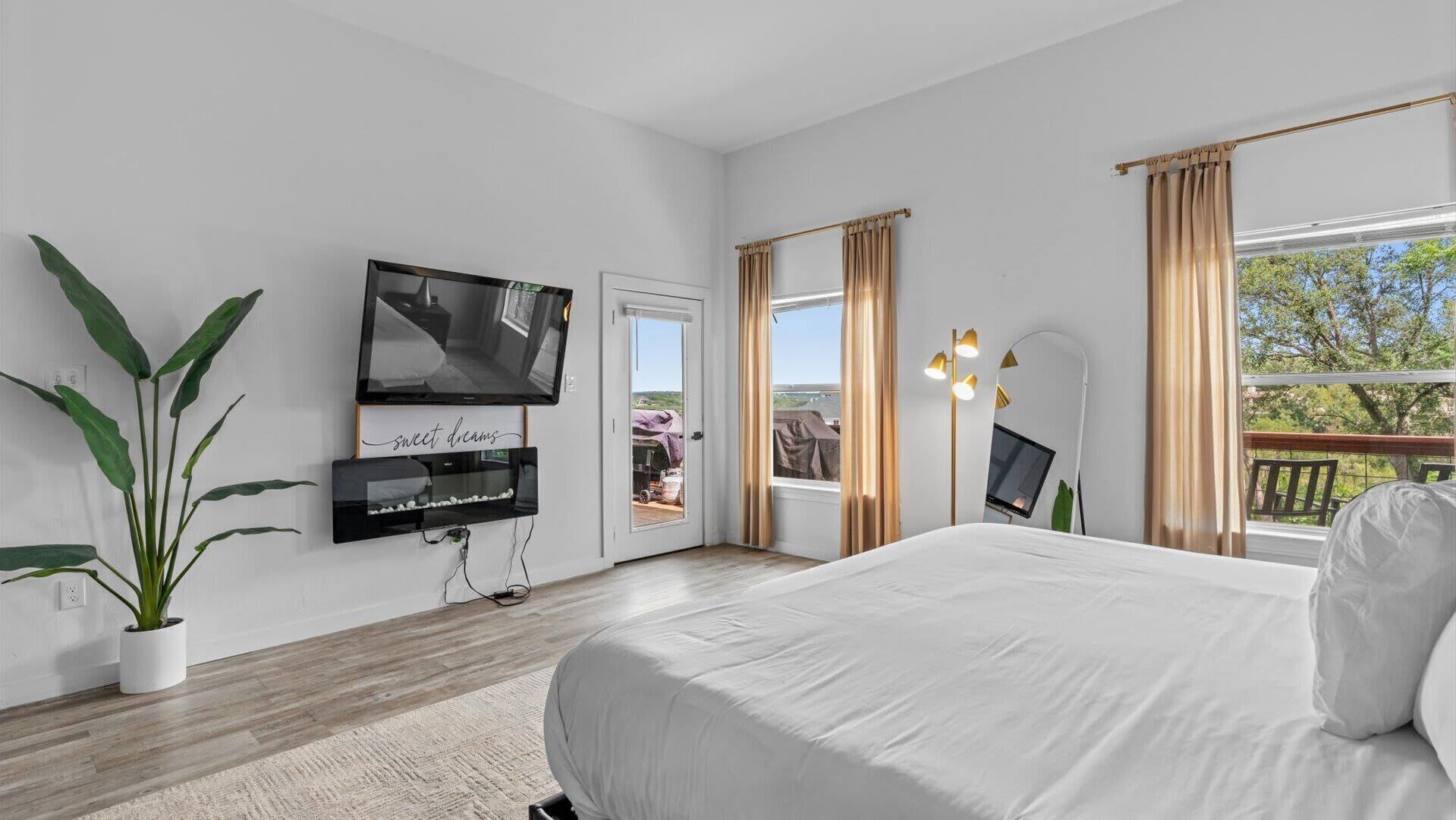A bedroom with a bed, a TV mounted on the wall, a potted plant, and a view of the outdoors.