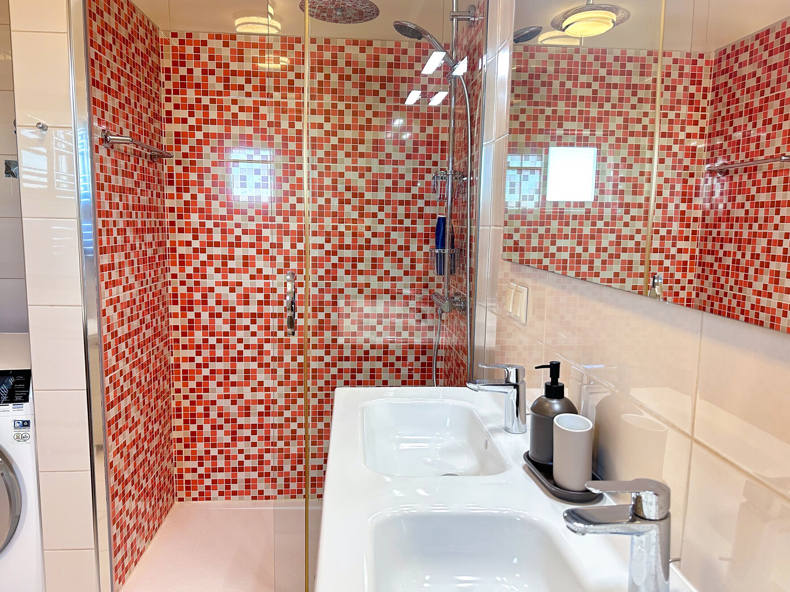 A modern bathroom with a glass shower enclosure, a white sink with a mirror, and a tiled wall with a red and white pattern.