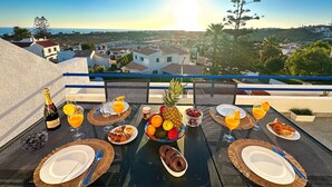 Een ontbijttafel met borden, bestek, een fles champagne en fruit op een balkon met uitzicht over een stadsbeeld.