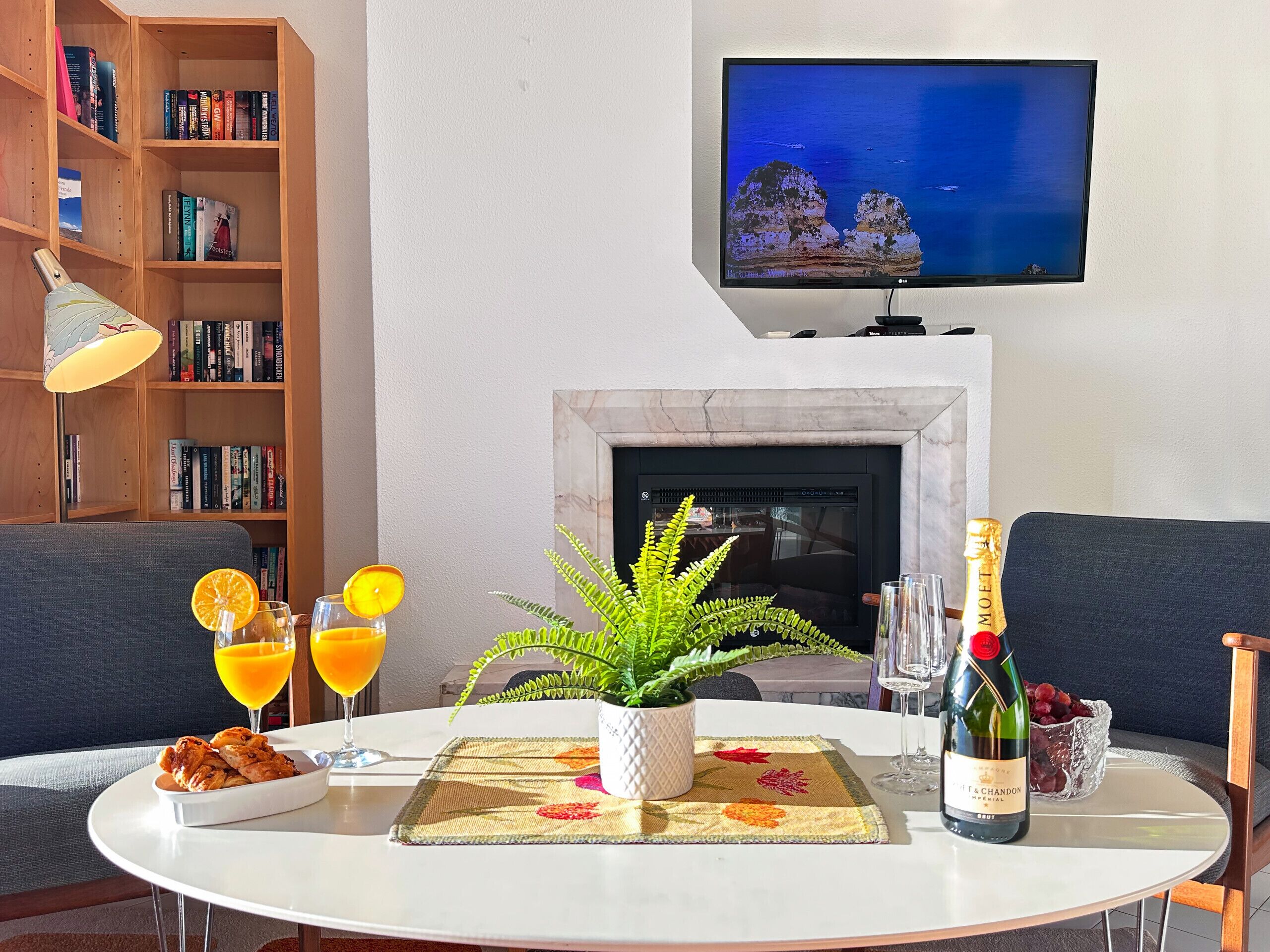 Un comedor con una mesa dispuesta para dos personas, un televisor sobre una chimenea y una estantería llena de libros.