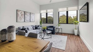 Un salon moderne avec un canapé, une table basse et une table à manger. De grandes fenêtres, un ventilateur de plafond et des œuvres d’art ornent les murs.