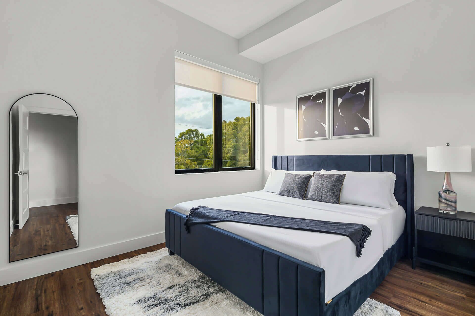 A modern bedroom with a large bed, a window with a view, a mirror, and a nightstand.