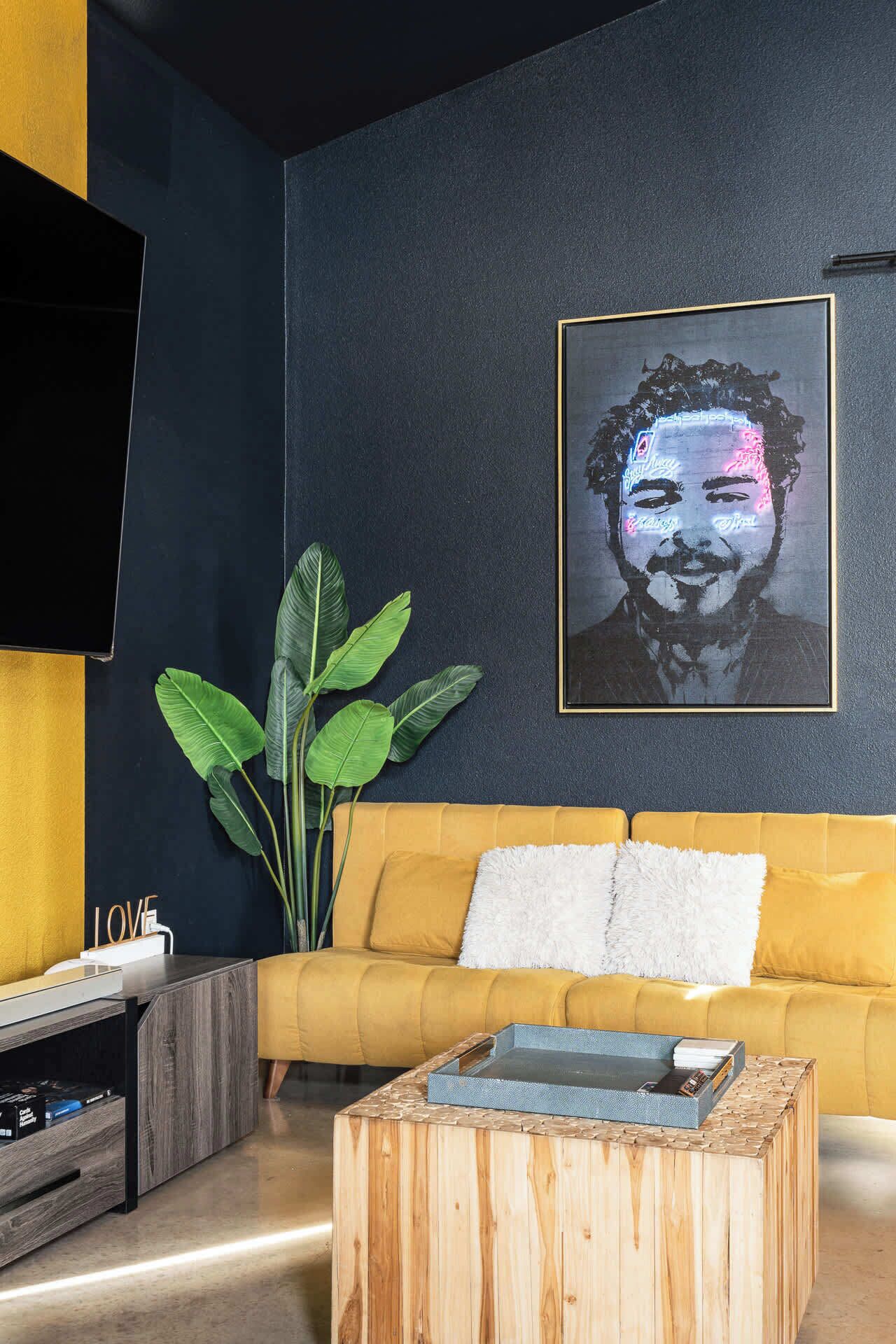 Una sala de estar moderna con una pared oscura, un sofá amarillo, una planta grande y una obra de arte enmarcada.