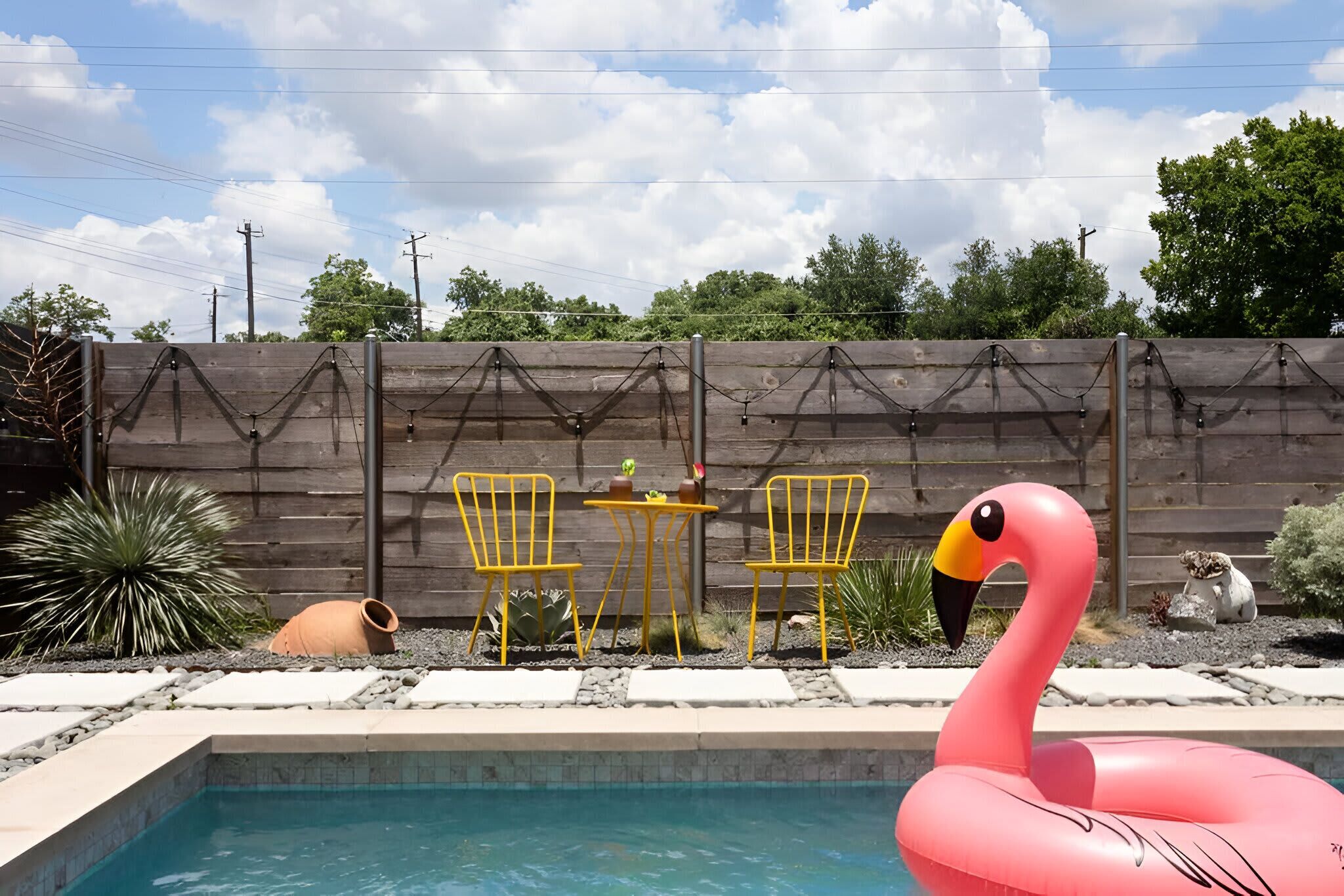 Une piscine agrémentée d’un flotteur en forme de flamant rose, entourée d’une clôture en bois, de chaises jaunes et d’une petite table sur laquelle est posée une plante en pot.