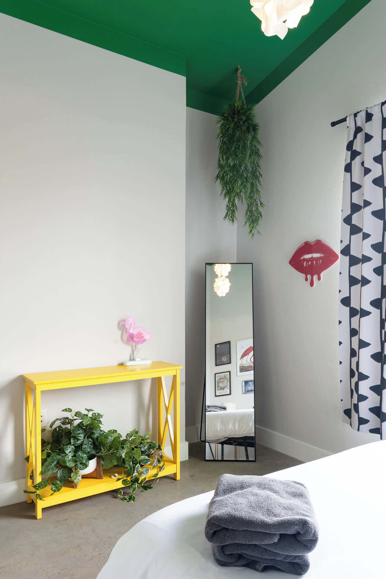 Une chambre à coucher avec un lit, une table de chevet ornée d’une plante, un miroir et une console jaune.