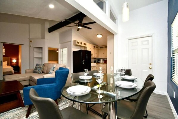 A modern living room with a glass dining table, blue armchairs, and a ceiling fan.