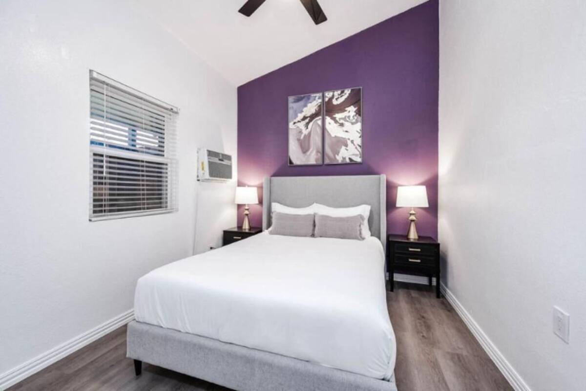 A bedroom with a bed, two bedside tables with lamps, a window with blinds, and a wall-mounted artwork.