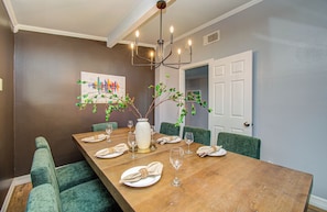 Une salle à manger avec une table en bois dressée pour six personnes, des chaises à l’assise rembourrée de tissu vert, un lustre et une porte donnant sur une autre pièce.