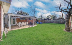 A backyard with a covered patio, a lawn, and a wooden fence. - Sleeps 16+ | Game Room + 75” TV | Fire Pit + BBQ (Houston)
