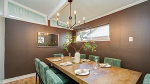 Une salle à manger avec une table en bois dressée pour six personnes, des chaises à l’assise verte, un lustre et un vase orné de branches.
