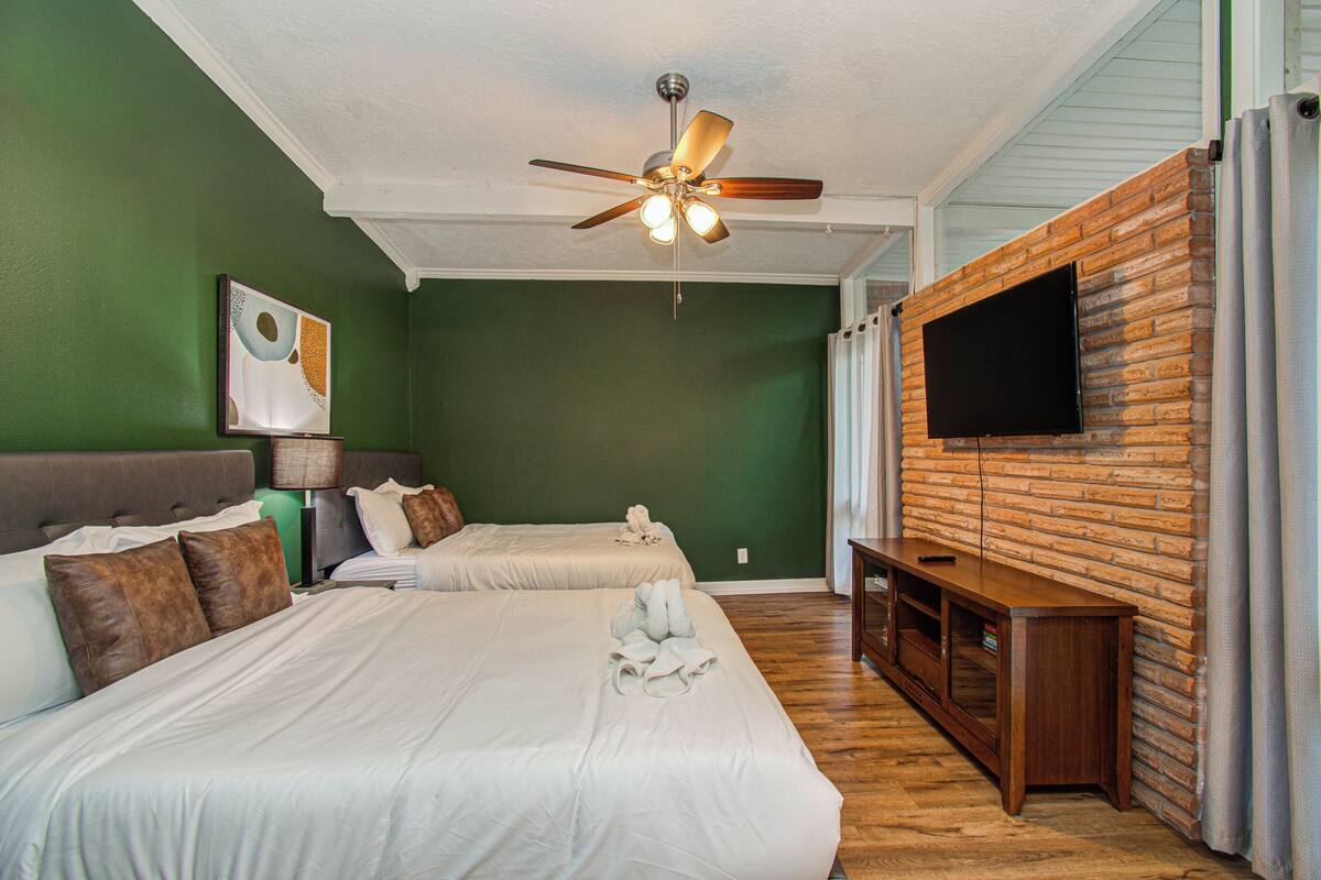 A hotel room with two beds, a ceiling fan, a flat-screen TV mounted on a brick wall, and a wooden bench.