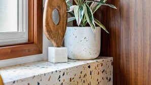 A corner of a room with a window, a wooden wall, a terrazzo countertop, a white speckled pot, and a wooden decorative piece.