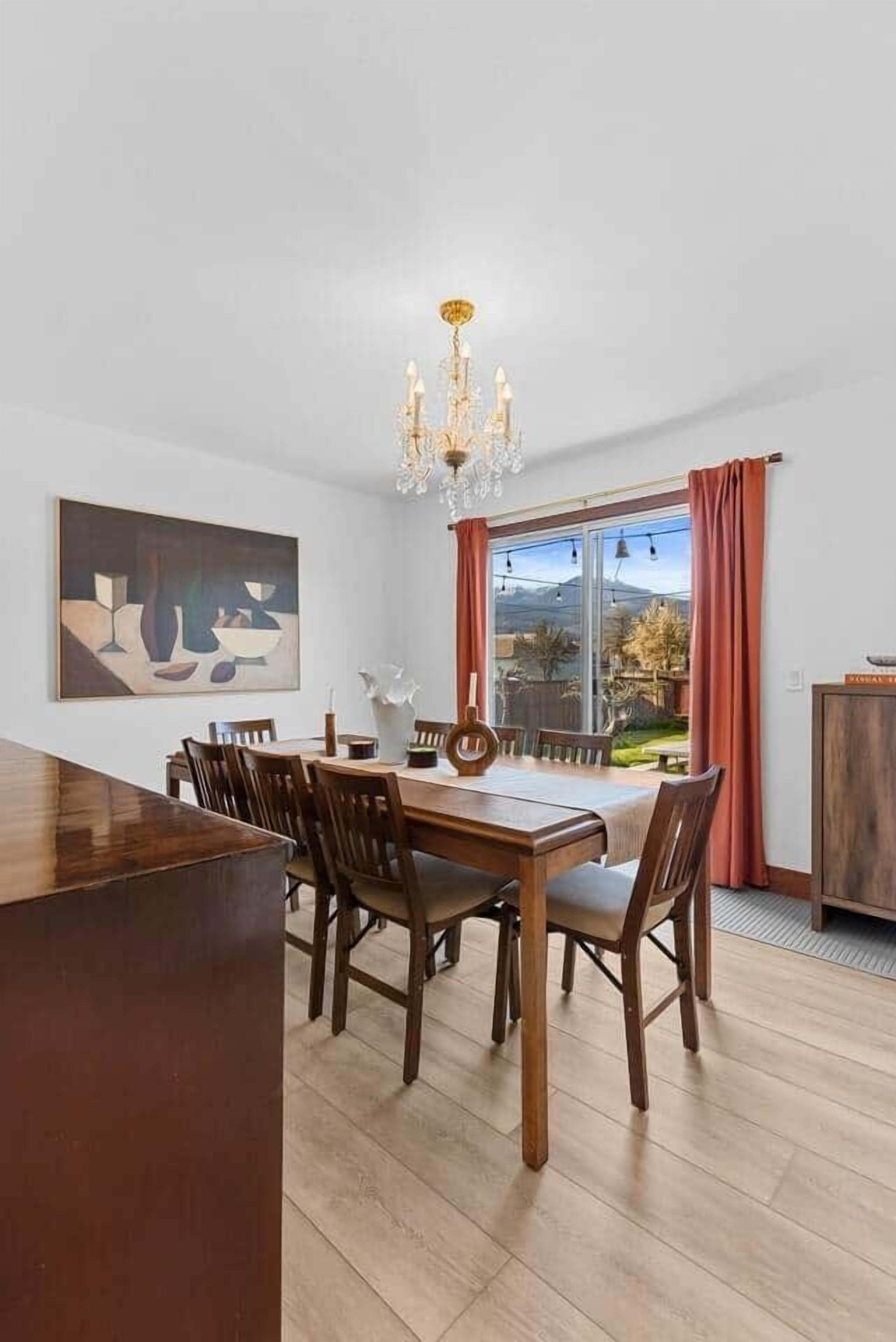 Une salle à manger avec une table et des chaises en bois, un lustre, un tableau au mur et une vue par une grande fenêtre.