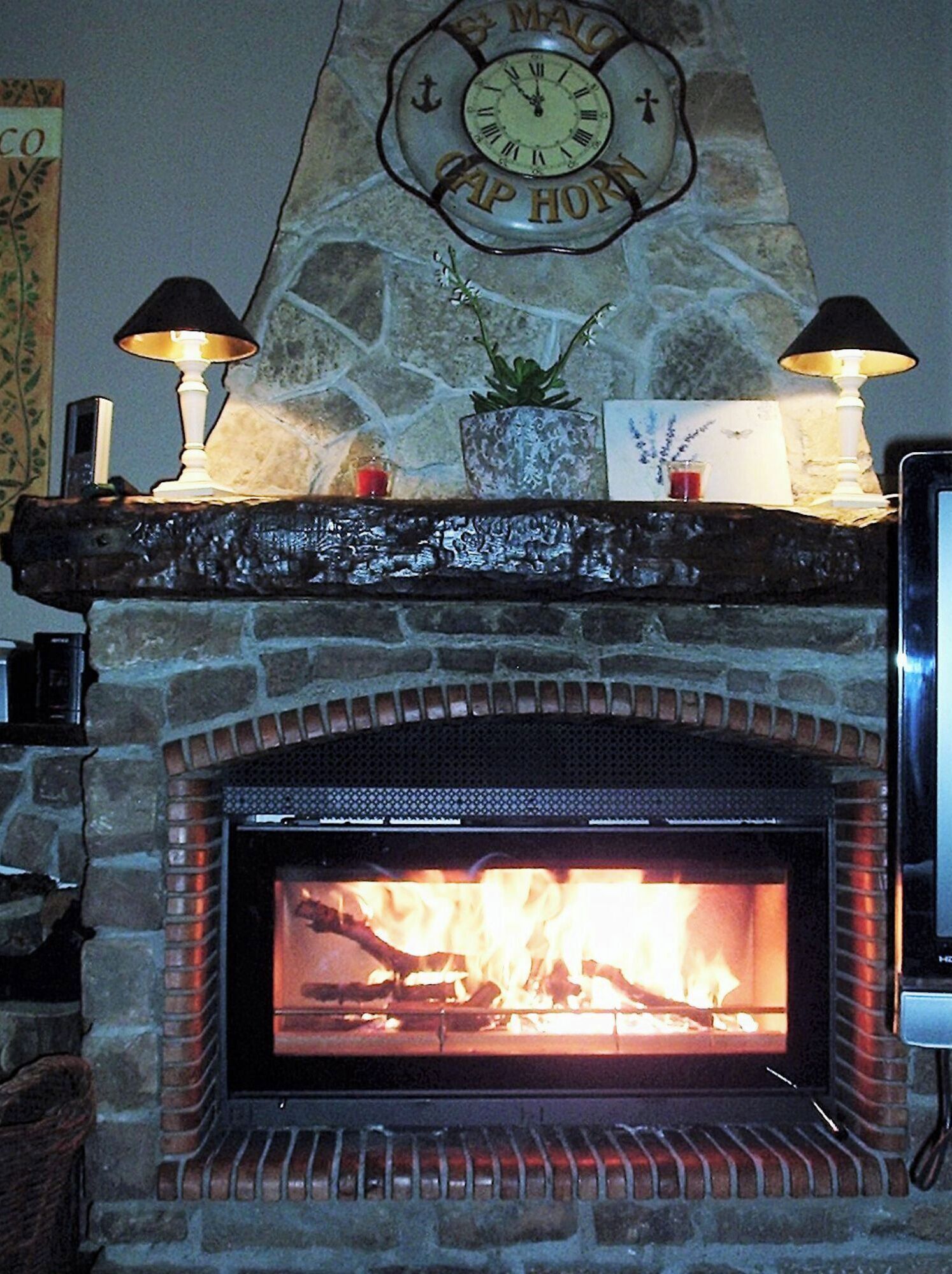 A stone fireplace with a clock above it, a plant on the mantel, and two lamps on either side.