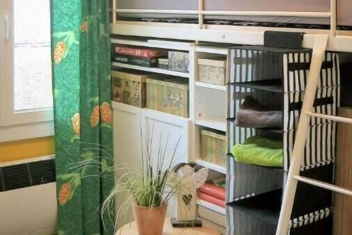 A bunk bed room with a small table and potted plant, a window with curtains, and a red cabinet.