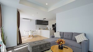 Ein modernes Wohnzimmer mit grauem Sofa, Hocker und rundem Couchtisch. Im Hintergrund ist eine Küchenzeile mit weißen Schränken und einer zentralen Kochinsel erkennbar.