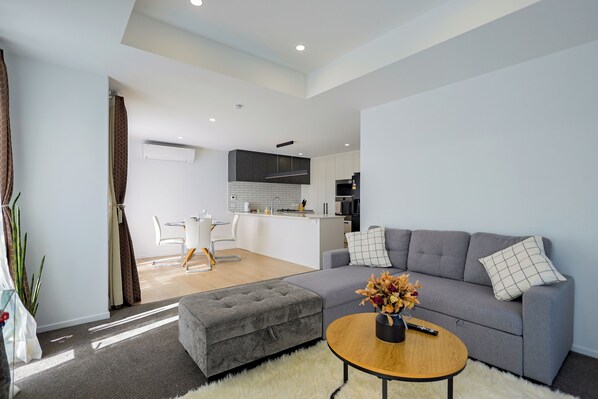 A modern living room with a grey sofa, ottoman, and round coffee table. There is a kitchen area in the background with white cabinets and a central island.