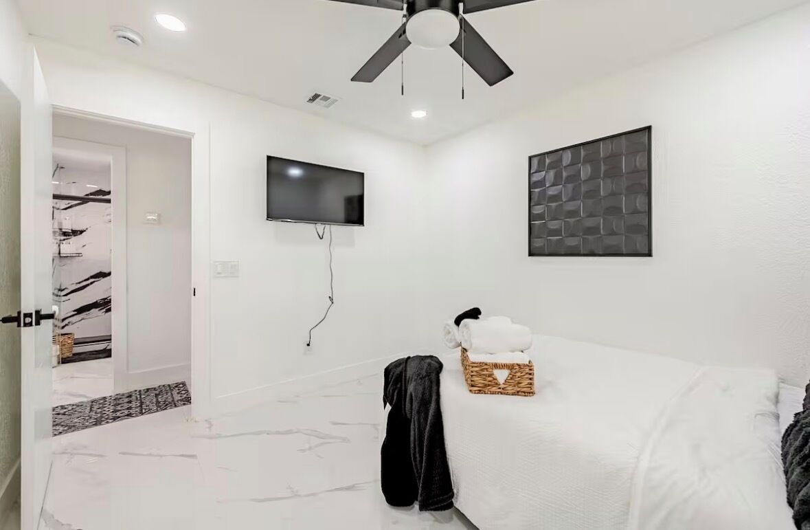 Ein modernes Hotelzimmer mit Deckenventilator, Flachbildfernseher, Wanddeko, einem Bett mit weißen Bettwäsche und einem Korbwäscheschrank.