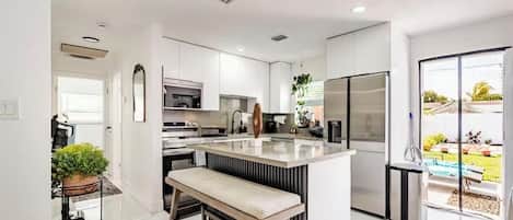 Une cuisine moderne avec des meubles blancs, un îlot central et une vue sur un jardin grâce à une porte-fenêtre coulissante.