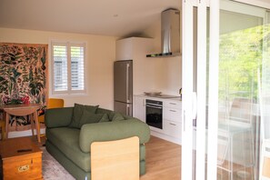 A compact living space with a sofa, a small dining table, a refrigerator, a stove, and a window with blinds. - City cottage oasis with sunken fire pit and private gated driveway parking. (Auckland)
