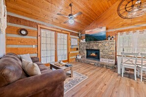 A cozy cabin interior with a stone fireplace, wooden walls, a ceiling fan, and a large window. - Perfect 2-bedroom cabin with AC in beautiful Suwanee (Sevier County)