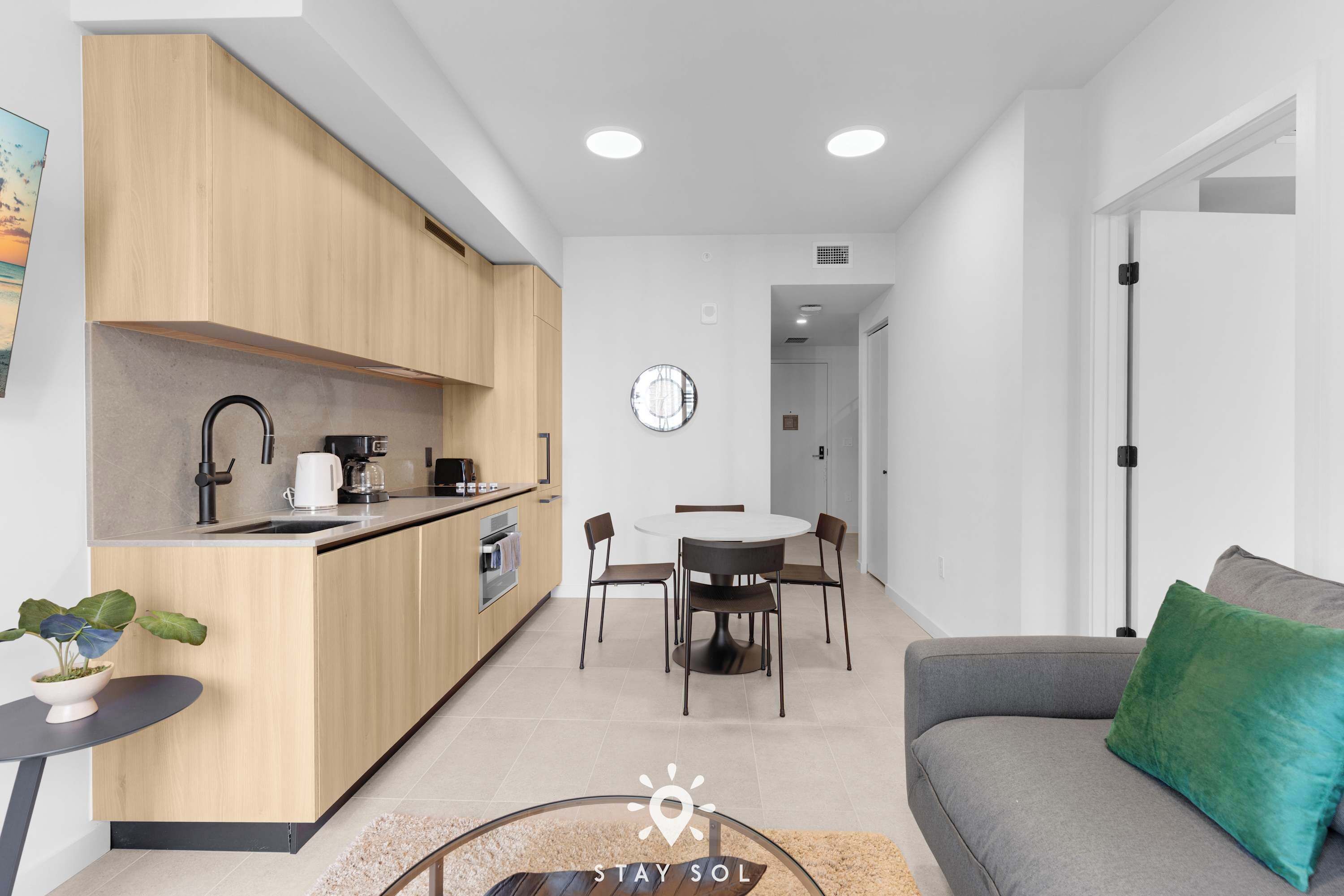 Une cuisine moderne avec des meubles en bois, une table à manger avec des chaises, un canapé gris avec un coussin vert et une petite table avec une plante.