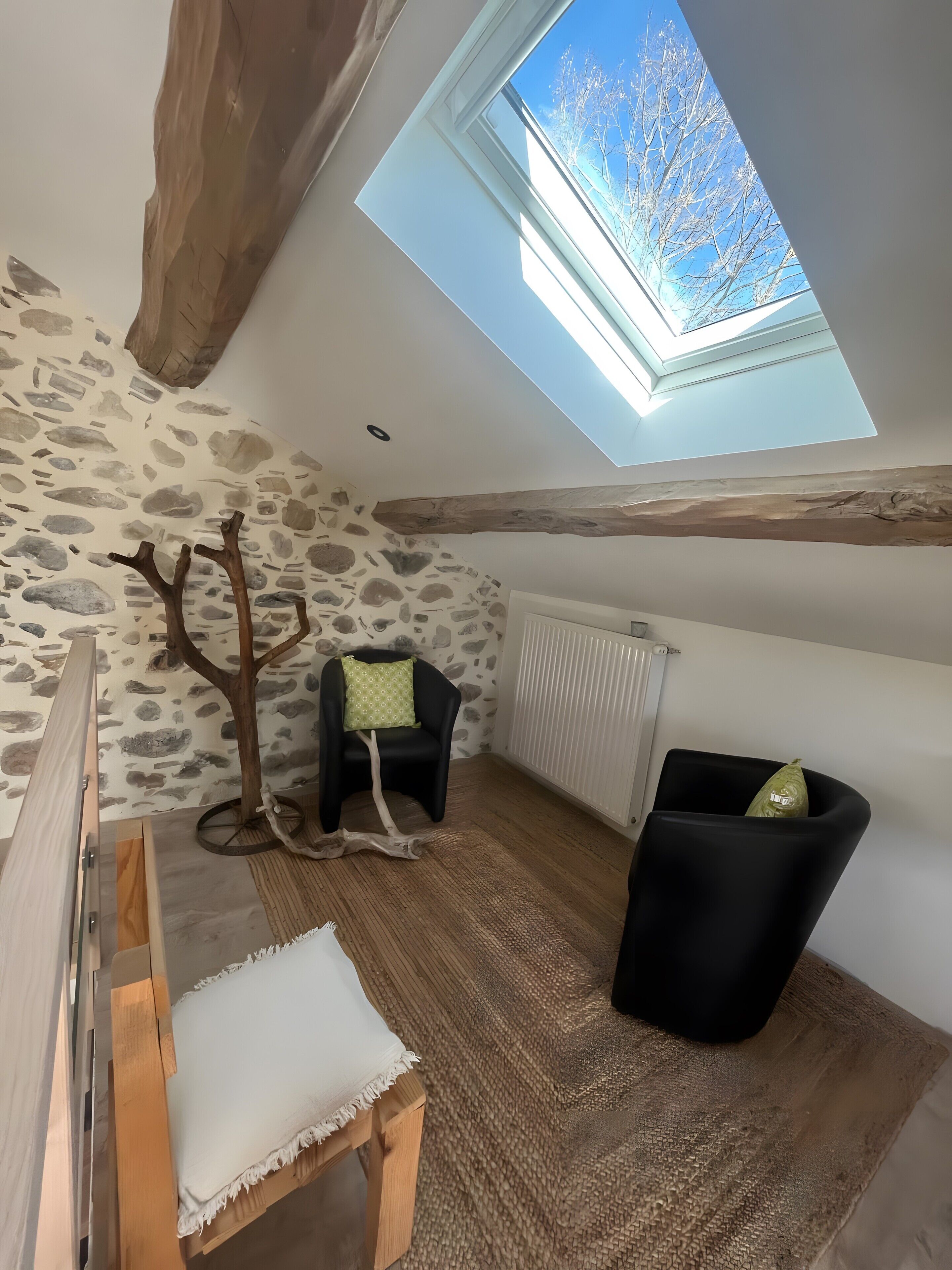Una stanza con soffitto a falde, parete in pietra, travi in legno, un velux, un termosifone, una panca in legno, una sedia nera e un tappeto intrecciato.