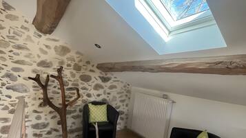 Una stanza con soffitto a falde, parete in pietra, travi in legno, un velux, un termosifone, una panca in legno, una sedia nera e un tappeto intrecciato.