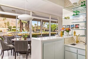 A modern kitchen with a large island, a dining area with chairs, and a view of a pool and mountains. - Palm Desert Mid-Century Modern | Private Spa, Pool, Piano & Walk to El Paseo (Palm Desert)