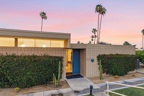 A modern building with a blue door, a well-maintained hedge, and palm trees in the background. - Palm Desert Mid-Century Modern | Private Spa, Pool, Piano & Walk to El Paseo (Palm Desert)