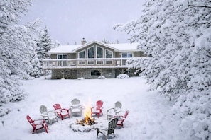 A snow-covered landscape with a house, a fire pit with chairs, and snow-laden trees. - Winter Escape | Sauna, Fireplace, Games (Parry Sound)