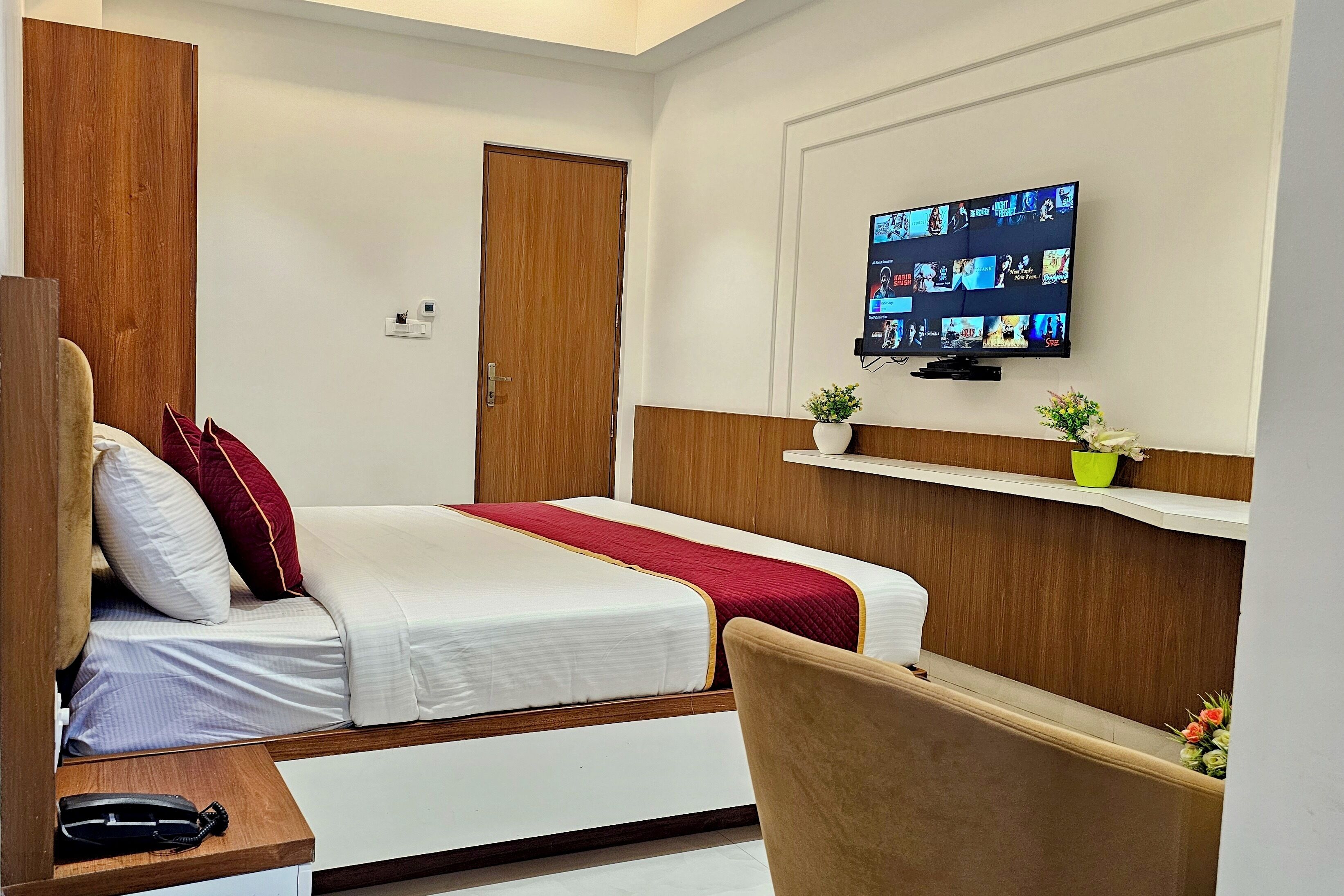 A hotel room with a bed, a TV mounted on the wall, a wooden wardrobe, a desk, and a chair.