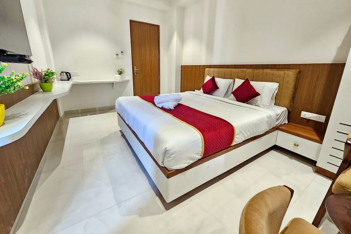 A modern hotel room with a large bed, a wooden headboard, a bedside table, a chair, and a small desk area.