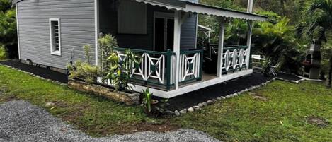A small, single-story house with a porch, surrounded by greenery and a gravel driveway.
