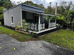 Ein kleines, eingeschossiges Haus mit Veranda, umgeben von Grün und einer Schotterzufahrt.