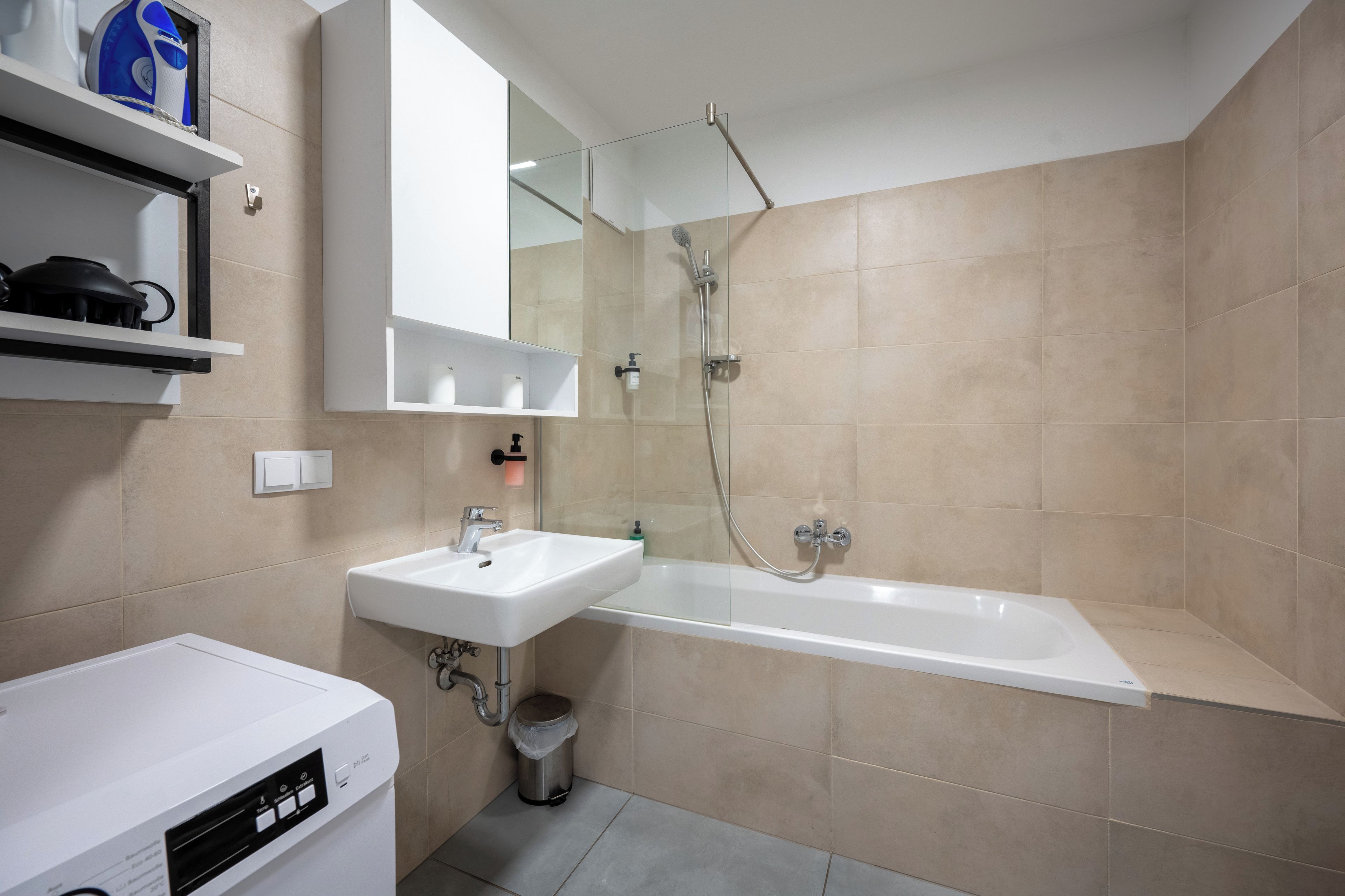 Un bagno moderno con lavandino bianco, vasca e doccia. Ci sono scaffali sopra il lavandino e un cestino della spazzatura vicino alla vasca.