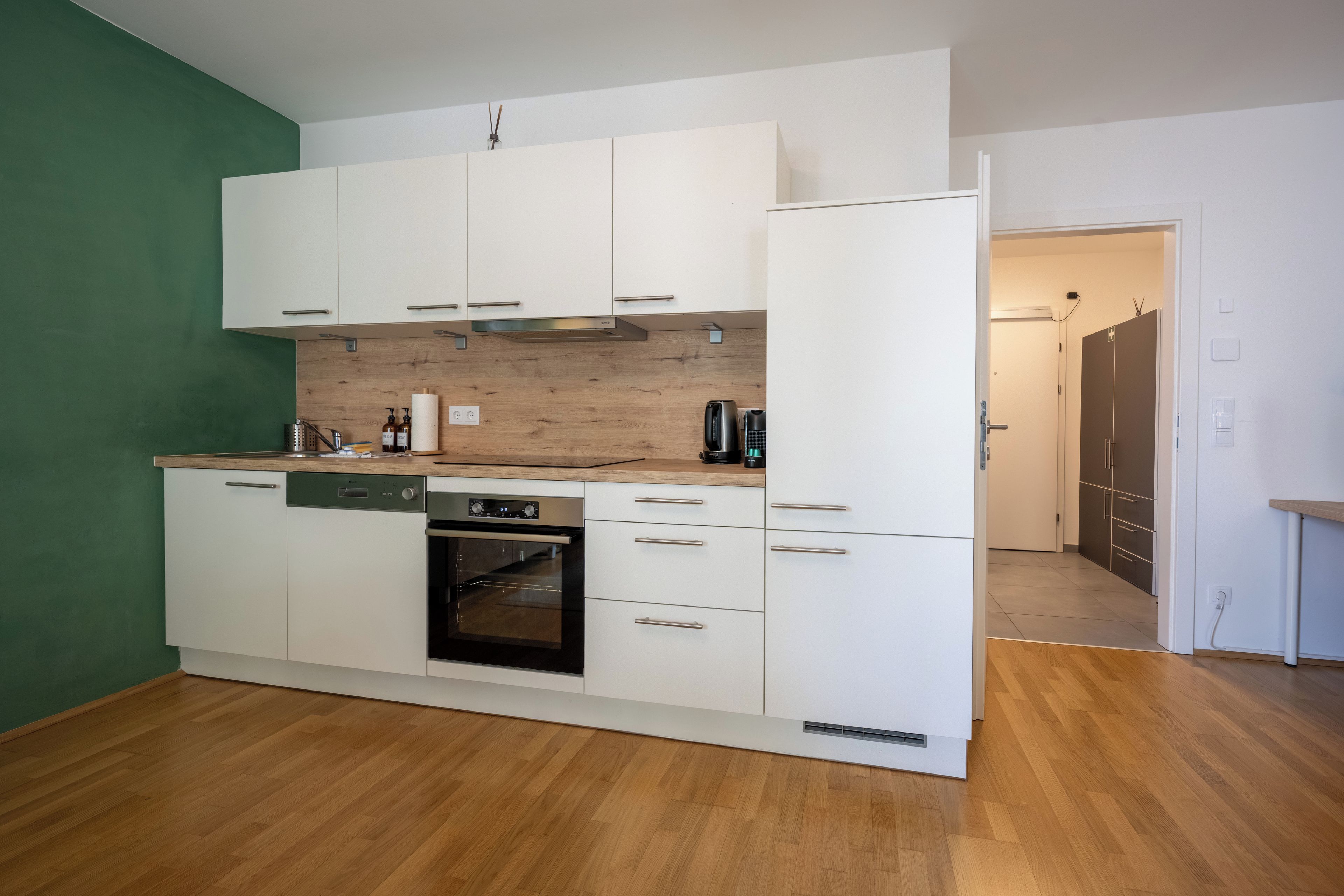 Een kleine keuken met witte kastjes, een inbouwoven en een houten spatwand.