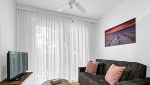 Een woonkamer met een donkergrijze bank, een ronde houten salontafel, een flatscreen televisie op een houten standaard, en een schilderij van een zonsondergang aan de muur.