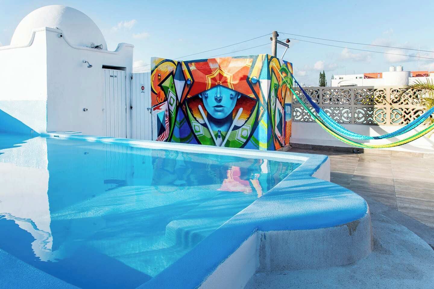 Une piscine sur le toit, agrémentée d’une fresque murale aux couleurs vives, un bâtiment blanc et un hamac.