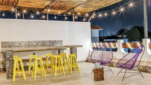 Eine Terrasse im Freien mit einem Holztisch, gelben Hocker, einem violetten Stuhl und Lichterketten.