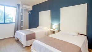 Ein Hotelzimmer mit zwei Betten, einem Nachttisch mit Lampe, einem Fenster mit Vorhängen und einer Klimaanlage.