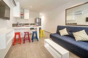 Ein modernes Wohnzimmer mit einem blauen Sofa, bunten Hocker, einem weißen Couchtisch und einer Küchenzeile mit weißen Schränken und einem Kühlschrank.
