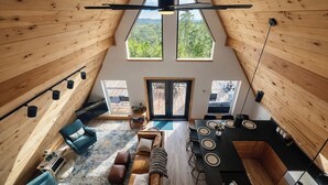 A high-ceilinged wooden interior with a dining area, seating, and a view through a large window. - Starfall A-Frame Hot tub + View + Firepit (Jasper)