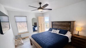 A bedroom with a bed, a ceiling fan, a nightstand with a lamp, a wooden bench, and a window with blinds. - Starfall A-Frame Hot tub + View + Firepit (Jasper)