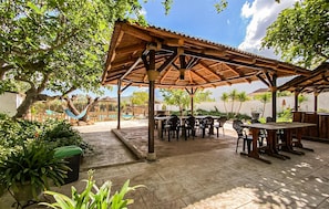 Een terras buiten met houten tafels en stoelen, omgeven door groen en een rieten dakconstructie.