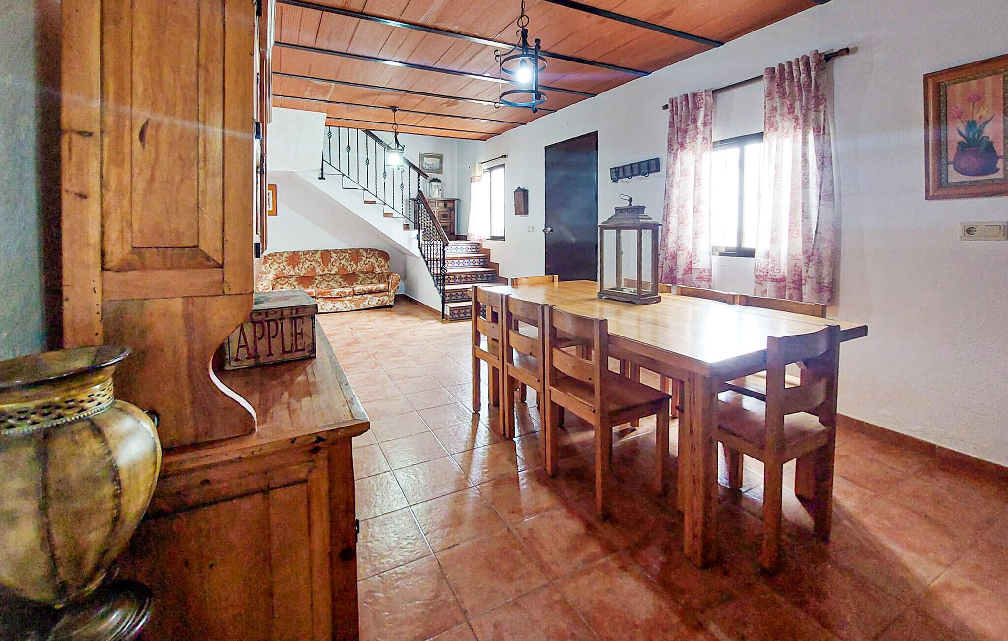 Un comedor con una mesa y sillas de madera, una escalera que sube a otro nivel y un gran ventanal con cortinas.