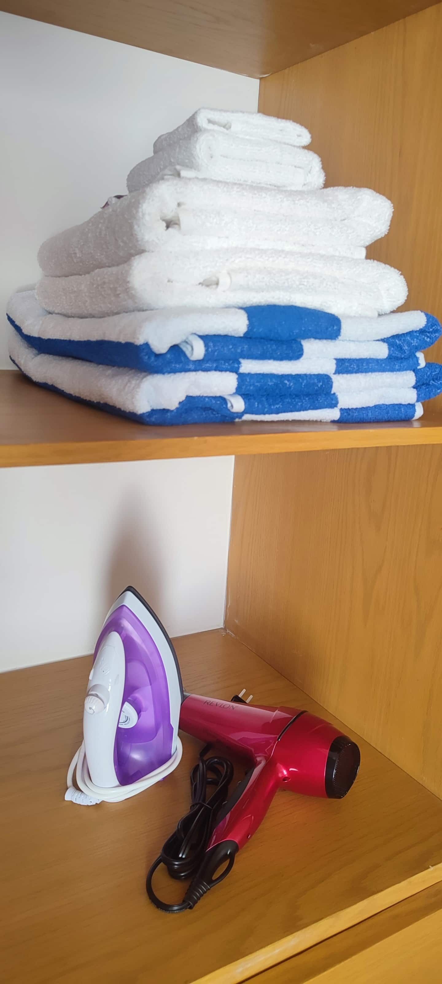 A stack of folded towels in white and blue on a shelf, a purple and white iron, and a red hairdryer on a wooden shelf.