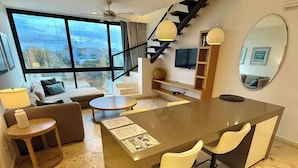 A modern kitchen with a central island, a dining area with chairs, a sofa, and a large window with a view. - Central 2 BR PH Gym, Private Rooftop, Garage, 406 (Playa del Carmen - Centro)